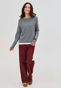Sivý pletený sveter s voľným strihom, vrstvený cez bielu košeľu, v kombinácii so širokými červenými nohavicami so vzorom čierneho leoparda. Hnedá obuv.