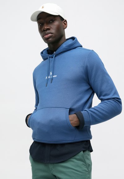 Νέος άνδρας που φοράει μπλε φούτερ με κουκούλα και τσέπη μπροστά, σκούρο πουκάμισο από μέσα, πράσινο παντελόνι και λευκό καπέλο μπέιζμπολ, στέκεται μπροστά σε λευκό φόντο.