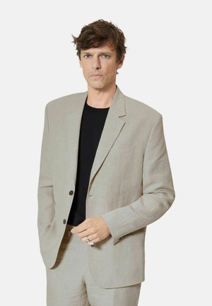 Mand med lyst brunt hår iført en beige jakkesæt og sort skjorte, stående med den ene hånd i lommen mod en ensfarvet baggrund.