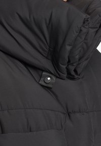 Gros plan d'une veste matelassée noire avec un col montant et un petit bouton-pression noir affichant un logo de feuille blanche.