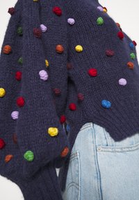 Pull en maille bleu marine orné de pompons colorés en rouge, orange, vert, violet et bleu. Côtés découpés et poignets côtelés.