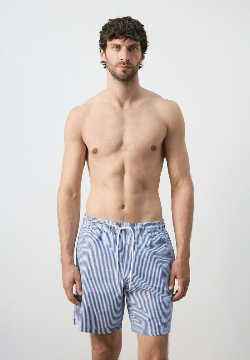 Mann mit dunklen lockigen Haaren, der blaue und weiße gestreifte Badeshorts trägt und vor einem einfarbigen hellen Hintergrund steht.