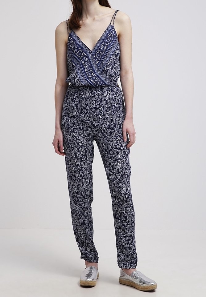 Navy blue jumpsuit featuring a patterned top with a crisscross design, thin straps, and tapered legs, paired with silver espadrilles.
