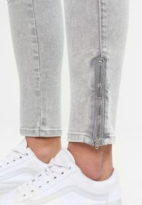 Pantalones de mezclilla gris claro con un detalle de cremallera plateada cerca del tobillo, combinados con zapatillas blancas con cordones y calcetines invisibles.