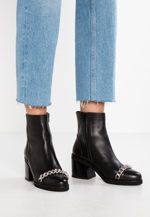 Botas de cuero negras hasta el tobillo con un tacón grueso y un detalle de cadena plateada en la parte delantera, combinadas con unos pantalones de mezclilla cortos en un tono azul claro.