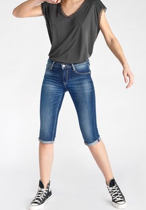 Mujer con pantalones capri de mezclilla azul remangados, top negro con escote en V y encaje, y zapatillas negras de caña alta, de pie con las piernas separadas.