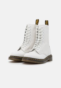 Botas de tornozelo brancas em couro com atacadores, com a biqueira redonda, costura amarela ao longo da sola e uma aba de puxar preta e amarela na parte de trás.