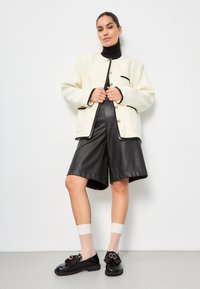 White knitted jacket with black trim and gold buttons, paired with black leather shorts. Black patent shoes with a bow accent and ankle socks.