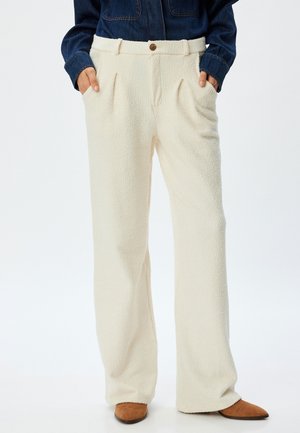 Pantalons en polaire couleur crème avec une surface texturée, des jambes larges et des plis sur le devant. Présente un système de fermeture avec bouton et des poches latérales.