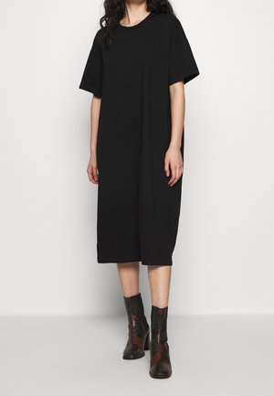 Femme portant une robe midi noire ample à manches courtes, associée à des bottines en cuir effet peau de serpent marron et noir, debout sur un fond blanc.