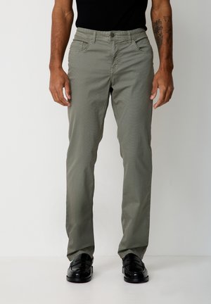 Man wearing olive green slim-fit pants, black t-shirt, black loafers, and a tattoo on his left forearm, standing on a white floor.