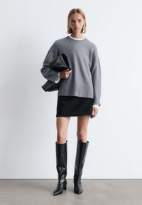 Grauer Strickpullover mit geripptem Saum, schwarzer Minirock und knielangen schwarzen Stiefeln mit Krokodilmuster. Das Model hält eine schwarze Jacke.