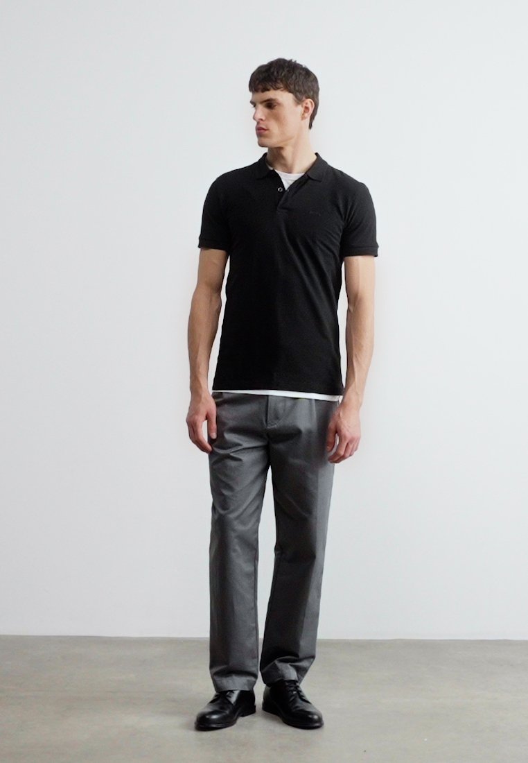 Jeune homme debout à l'intérieur, portant un polo noir, un pantalon gris et des chaussures noires, regardant de côté contre un mur blanc uni.
