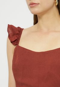 Γυναίκα που φορά κορμάκι χωρίς μανίκια σε σκουριά απόχρωση με βολάν στις τιράντες και χρυσό κρίκο, δείχνοντας τον λαιμό και τα χείλη.