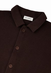 Bruine geribbelde polo met een zachte textuur, voorzien van een kraag, drie bruine knopen en een label met de tekst "OH APRIL."