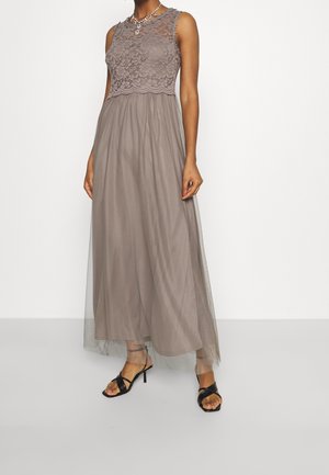 Bezrękawowa taupe sukienka z koronkowym gorsetem i przezroczystą spódnicą nałożoną na warstwy, w połączeniu z czarnymi sandałami na obcasie z paskami, na stojącej kobiecie.