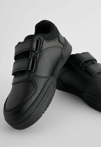 Une paire de chaussures de sport noires avec deux bandes velcro et des semelles texturées sur un arrière-plan gris clair.