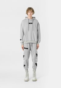 Sudadera gris claro con una etiqueta negra, combinada con pantalones jogger a juego que presentan patrones de alas en blanco y negro, y botas de combate blancas.