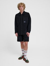 Jonge man met krullend blond haar die een zwarte jas met rits draagt, zwarte korte broek, witte gestreepte sokken en groene sneakers, staand op een witte achtergrond.