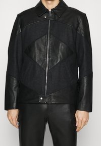 Veste en cuir noir et en denim avec un design de panneaux géométriques, col droit, zip frontal et fermeture à pression à la taille. Texture lisse.