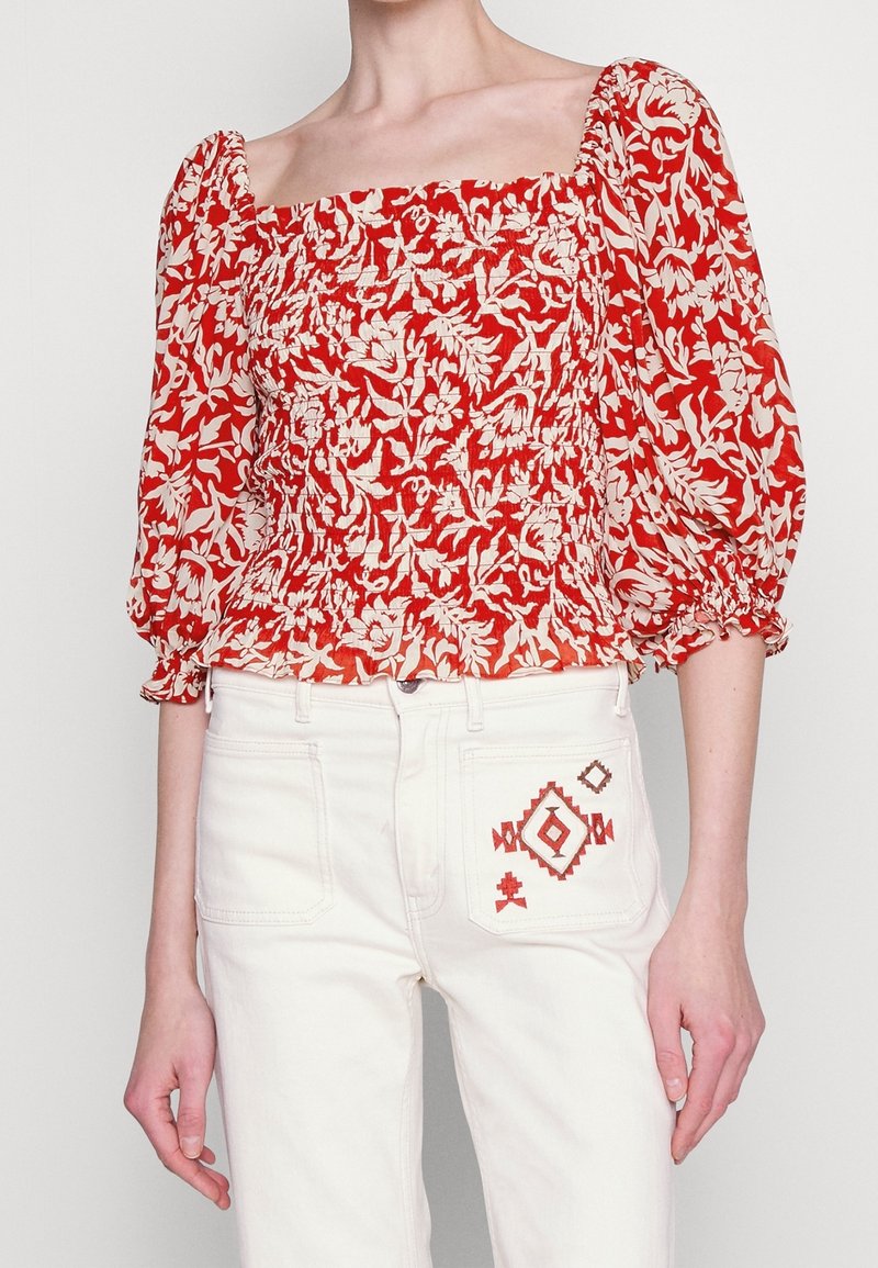 Femme portant un chemisier smocké rouge à fleurs avec manches bouffantes et un pantalon blanc orné d'une broderie géométrique rouge sur la poche arrière.