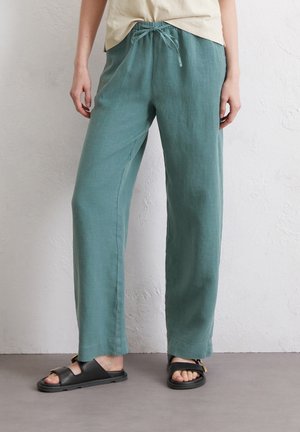 Persona con pantalones anchos de color azul verdoso con cintura ajustable y sandalias negras de punta abierta, de pie contra una pared blanca.