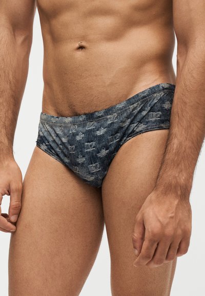 Buste et cuisses d'un homme portant un maillot de bain style slip à motif foncé, bras détendus le long du corps sur fond uni.
