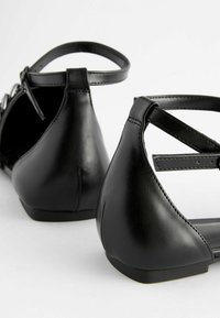 Bailarinas de cuero negro con un tacón bajo, que presentan un diseño con recortes y correas para el tobillo ajustables. Textura suave y detalles de costura mínimos.