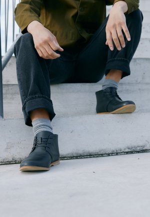 Stivali ankle in pelle nera con finitura liscia, punta rotonda e design con lacci. La suola è marrone chiaro e offre un accento di contrasto.