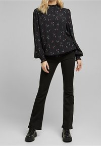 Schwarze Bluse mit floralem Druck in Rosa und Grün, hat gepolsterte Ärmel und einen hohen Ausschnitt. Kombiniert mit schwarzen, weit ausgestellten Hosen und klobigen schwarzen Stiefeln.