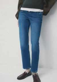 Osoba ubrana w niebieskie spodnie typu straight-leg, beżowe skarpetki, ciemnobrązowe mokasyny, warstwową, kraciastą kurtkę oraz ciemny dzianinowy sweter z białym wykończeniem.