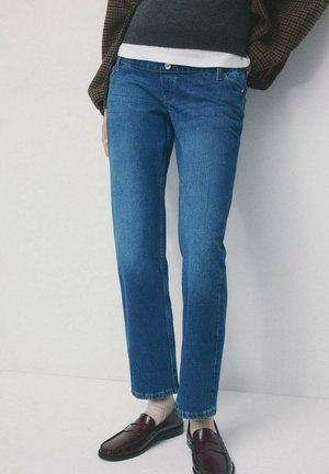 Osoba ubrana w niebieskie spodnie typu straight-leg, beżowe skarpetki, ciemnobrązowe mokasyny, warstwową, kraciastą kurtkę oraz ciemny dzianinowy sweter z białym wykończeniem.