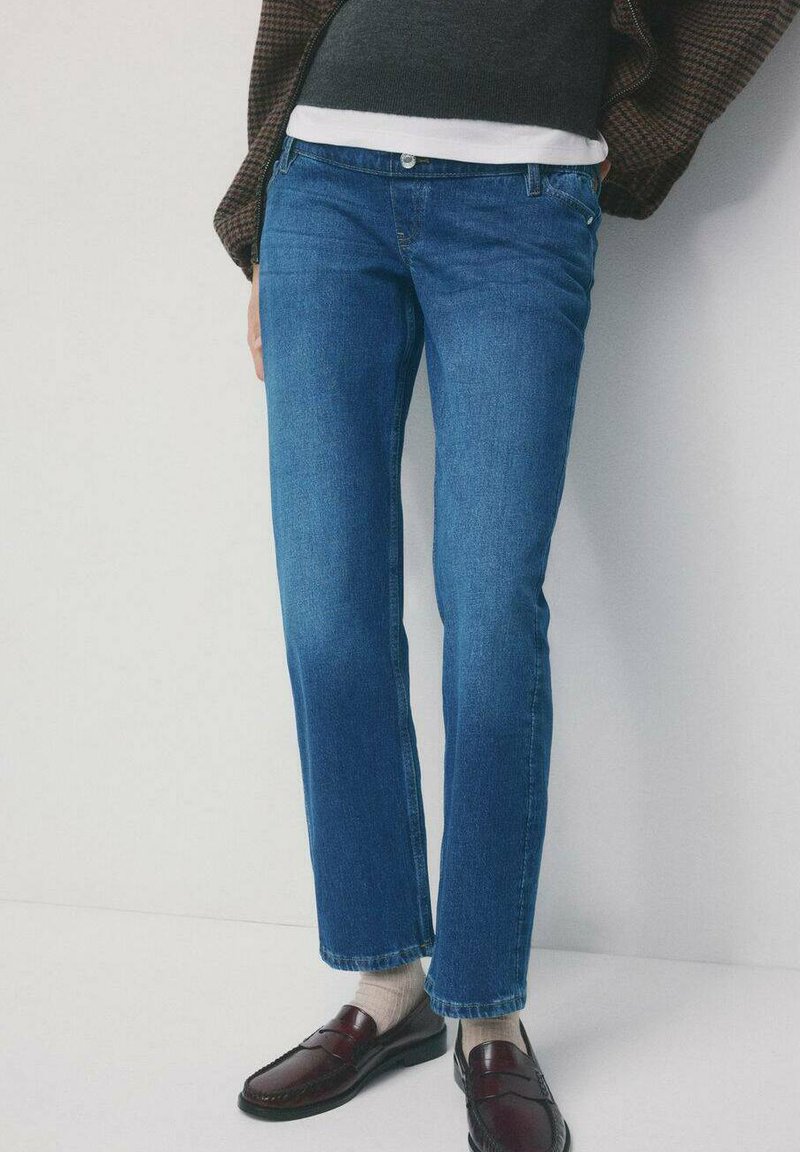 Osoba ubrana w niebieskie spodnie typu straight-leg, beżowe skarpetki, ciemnobrązowe mokasyny, warstwową, kraciastą kurtkę oraz ciemny dzianinowy sweter z białym wykończeniem.