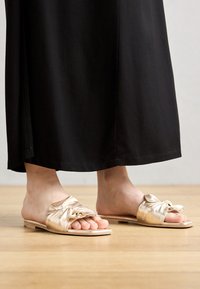 Sandales ouvertes en satin doré avec un accent noué sur le dessus, un design à bout ouvert et une semelle plate en bois. Portées avec un vêtement noir.