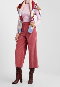 Pinkfarbener Rollkragenpullover kombiniert mit einer mehrfarbigen Strickweste, pinkfarbenen Cordhose mit weitem Bein und glänzenden roten Stiefeletten mit Schlangenprint.