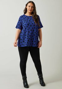 Blå kortærmet top med sorte prikker, tekstureret stof, rund hals; kombineret med sorte skinny bukser og knæhøje stilettoboots.
