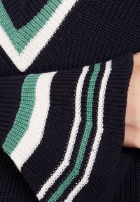 Námořnický pletený svetr s výstřihem do V, s kontrastními pruhy v zelené, bílé a černé barvě na rukávech a lemu. Texturovaný vzor.