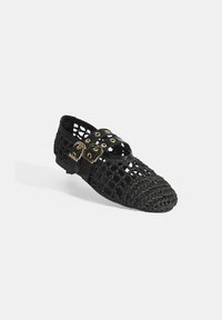 Schwarzer gewebter Schuh mit offenem Design, akzentuiert durch zwei verstellbare Riemen mit goldenen Beschlägen. Die Sohle ist flach und strukturiert.