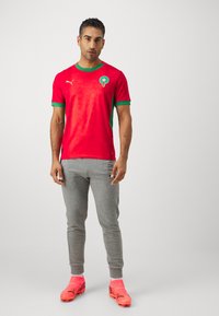 Maglietta da calcio rossa con accenti verdi e emblema marocchino, abbinata a pantaloni della tuta grigi e scarpe da calcio fucsia vivaci. La texture presenta un motivo a stelle.