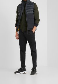 Gepolsterte schwarze Weste über einer grünen Jacke mit Reißverschluss, kombiniert mit schwarzen, zulaufenden Hosen und schwarzen Turnschuhen mit weißen Akzenten.