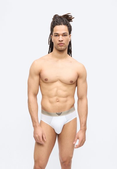 Μυώδης άνδρας με ντρέντλοκ στέκεται προσανατολισμένος προς τα εμπρός, φορώντας λευκά εσώρουχα με μπεζ ζώνη στη μέση, πάνω σε απλό φωτεινό φόντο.