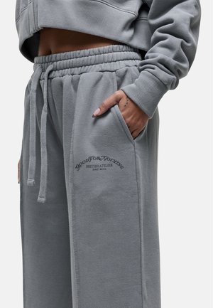 Pantaloni tuta grigi con cintura elastica, tasche laterali e dettagli del logo ricamati. Tessuto morbido con vestibilità rilassata e cuciture visibili.