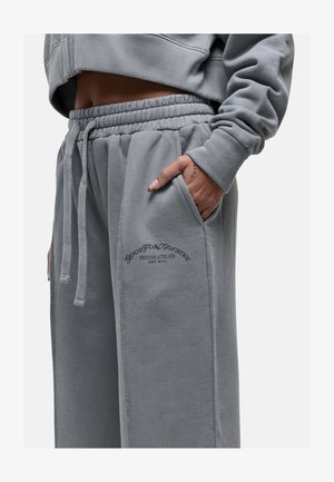 Pantaloni tuta grigi con cintura elastica, tasche laterali e dettagli del logo ricamati. Tessuto morbido con vestibilità rilassata e cuciture visibili.