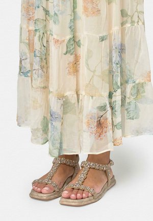 Picioare încălțate cu sandale plate aurii cu bretele în formă de T împodobite cu bijuterii, asortate cu o fustă lungă, transparentă cu imprimeu floral în culori pastelate delicate.