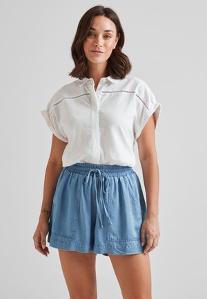 White linen shirt with short rolled sleeves and button closure, paired with light blue denim shorts featuring an elastic waistband and side pockets.