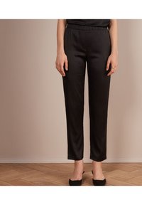 Svarta satinbyxor med elastisk midjeband, smalnade ben och slät textur, tillsammans med svarta klackskor, mot en ljus bakgrund.