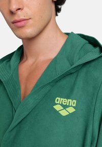 Accappatoio verde in cotone con cappuccio e colletto a coste. Presenta un logo giallo "arena" sul petto. Tessuto liscio, design aderente.