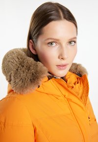 Oranžová péřová bunda s měkkou béžovou kožešinou podšitou kapucí, s hladkým povrchem a viditelnými švy na těle.