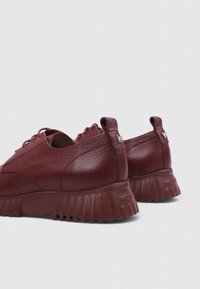 Un par de zapatos de cuero color rojo oscuro con cordones, suelas de goma gruesa texturizada y pestañas en el talón, mostrados desde un ángulo lateral trasero.