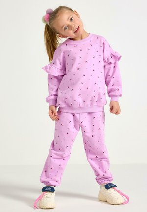 Młoda dziewczyna z blond kucykiem, ubrana w różową, kwiatową bluzę z falbankami i spodnie, pozująca z przechyloną głową i zaciśniętymi dłońmi.